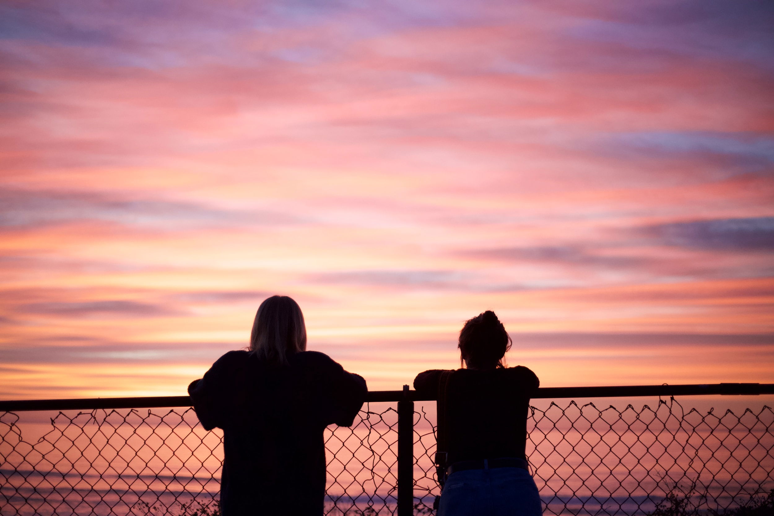 A pair of friends side by side, facing away from the camera while leaning on a fence, enjoying the sunset together.