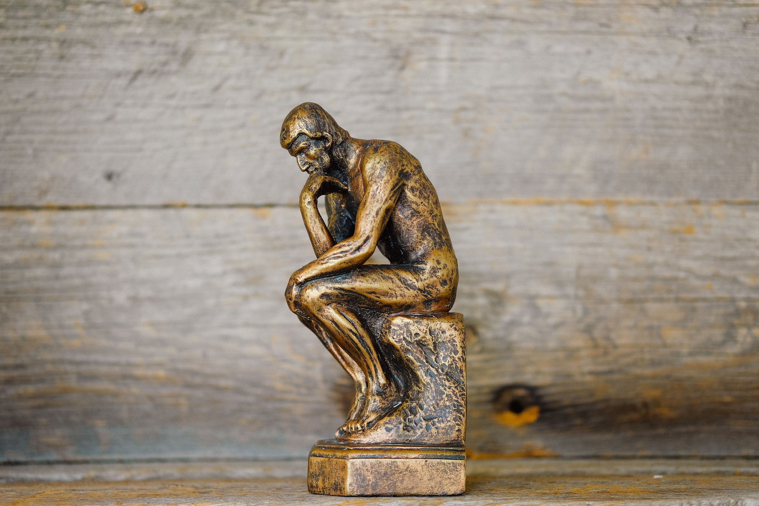 A small statue of The Thinker sculpture by Auguste Rodin, placed atop a table in front of a wooden plank background.