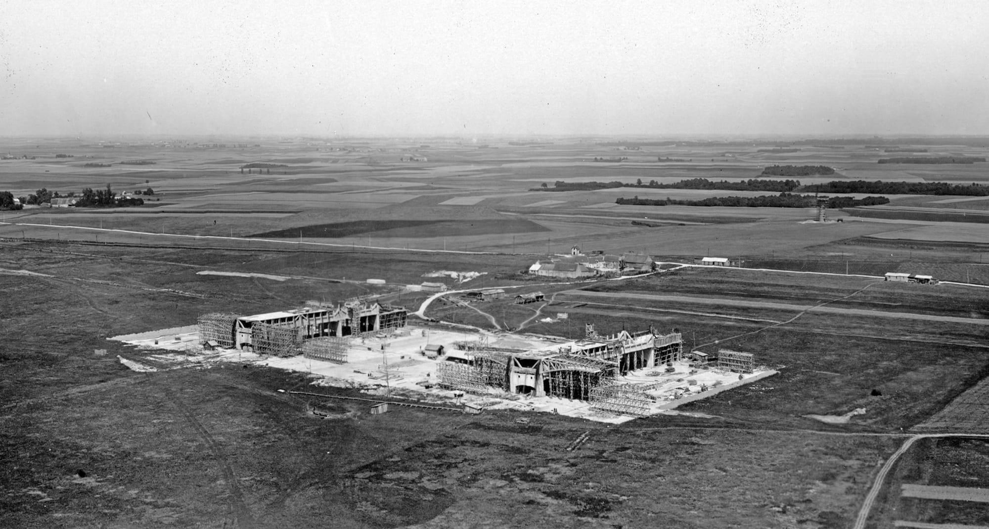 Vue aérienne du chantier des deux premiers hangars Caquot. On remarque en arrière plan la ferme du Coudray qui servira de locaux pour les personnels des Ponts et Chausées, au fond le château d’eau en construction juste devant le bois d’eau. Cette photo est certainement prise vers l’été 1933. (Photo USID/Bricy)