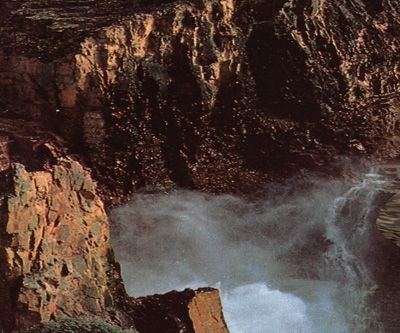 This image shows a white, frothy stream flowing down a canyon between tall, red, volcanic cliffs.