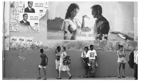 People, dressed in Western clothing, stand in front of a mural depicting the importance of safe sex, another Western import.