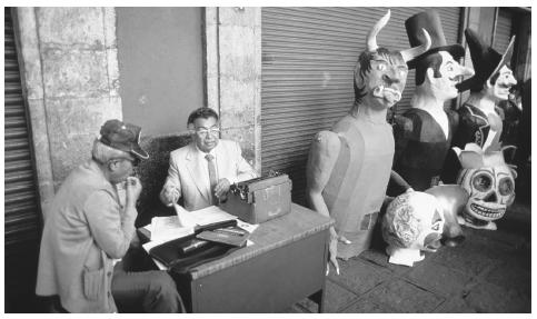 A scribe works with a client on a sidewalk in Mexico City. Beside them are papier-mâché figures for a Holy Week festival.