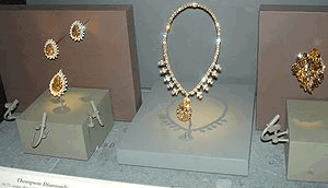 A museum display of jewelry items. Three brooches each consist of a large brown central gem surrounded by many clear small stones. A necklace has a large brown gem at its bottom and its string is all covered with small clear gems. A cluster-shaped decoration contains many brown gems.