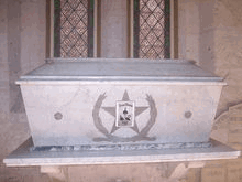 A white marble coffin sits on a ledge in front of stained glass windows. On the front of the coffin is a large 5-pointed star. Engraved within the star are the words 