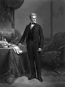 Man stands in white shirt and black pants and coat with right hand on desk and left hand at his side.