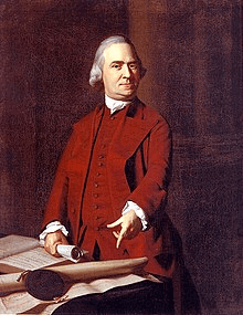 A stern middle-aged man with gray hair and wearing a dark red suit. He is standing behind a table, holding a rolled-up document in one hand, and pointing with the other hand to a large document on the table.