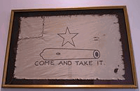 A white flag, with a five-pointed-star over the outline of a small cannon, with the words Come and Take It below. The left corner of the flag has been torn off and sewn back on.