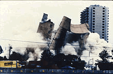 The Alfred P. Murrah building is being demolished, and the image shows the building in mid-collapse. A Ryder truck is visible at the bottom left, and the Regency Towers building can be seen in the background at the far right. The demolition has created large clouds of dust that take up a portion of the image.