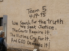 A woman, at the left of the image, is reading a black spray paint message written on a brick wall. The message reads 