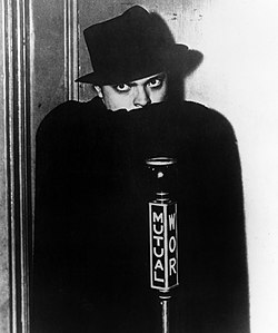 Man in black hat concealing the bottom of his face with a black cape and gazing fiercely. A microphone in front bears the word 