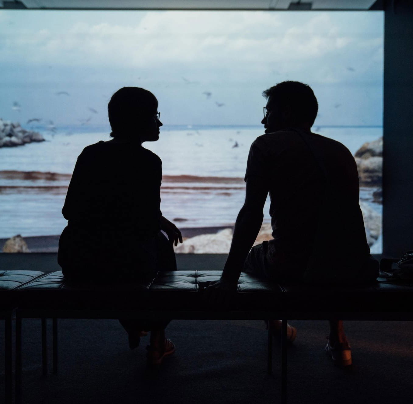 A silhouette of two people talking on a bench with a view of a bay.